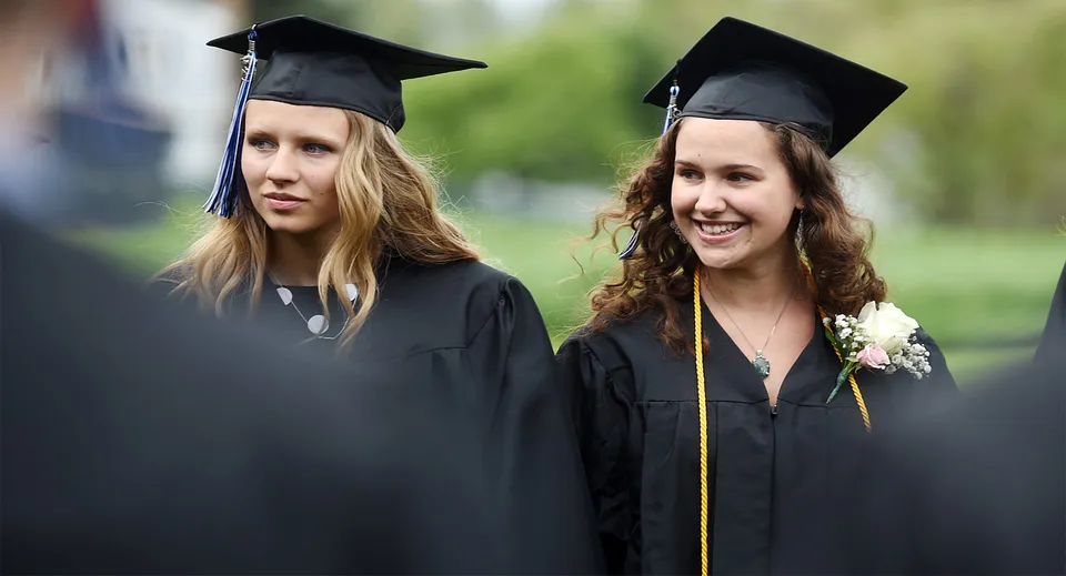 Due diplomande della Stillwater Christian School posano insieme con la toga da diploma su uno sfondo verde.