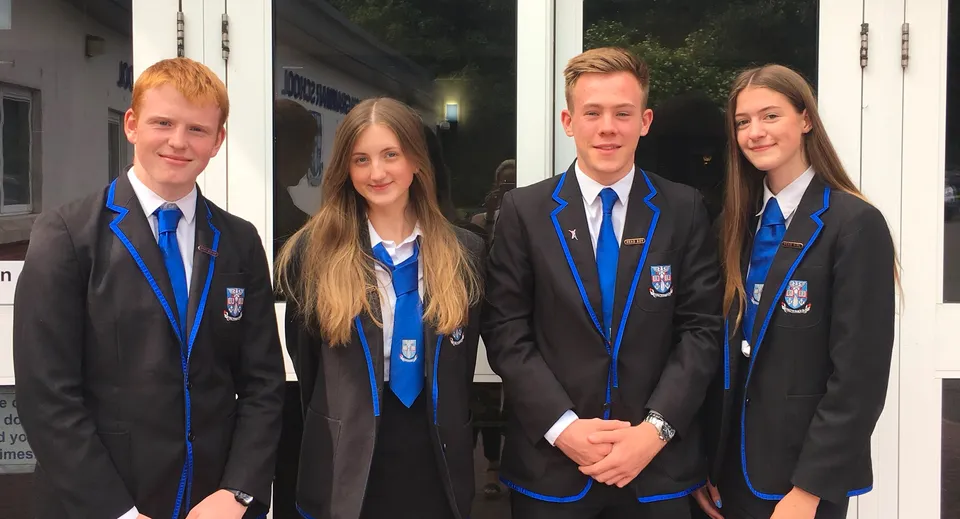 Quattro studenti della Musselburgh Grammar School in divisa scolastica sono in piedi davanti a un edificio di notte.