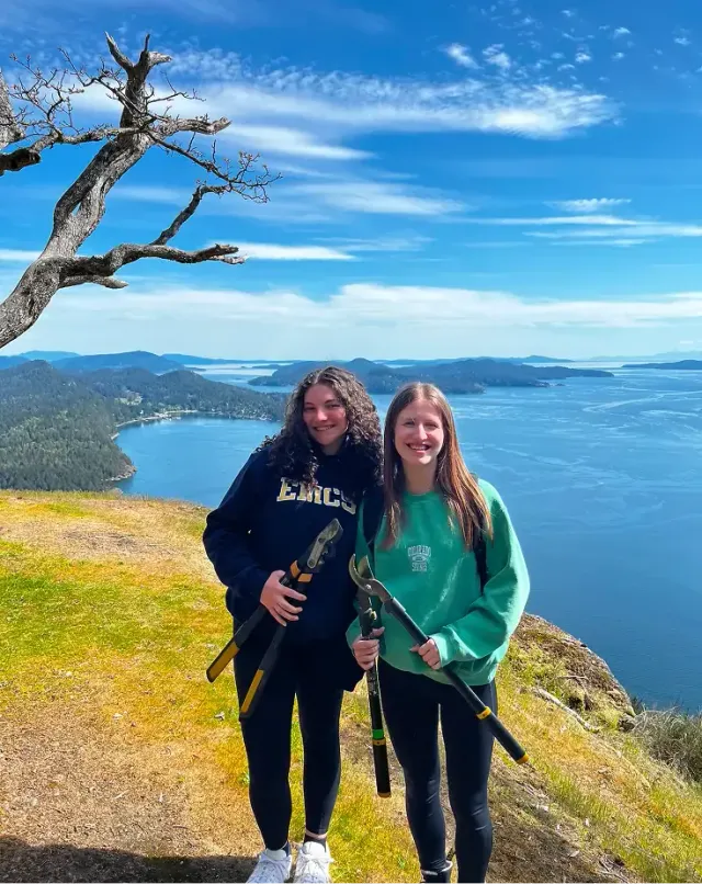 Due studentesse sorridenti su una collina davanti al mare, con cesoie in mano, durante uno scambio in Canada