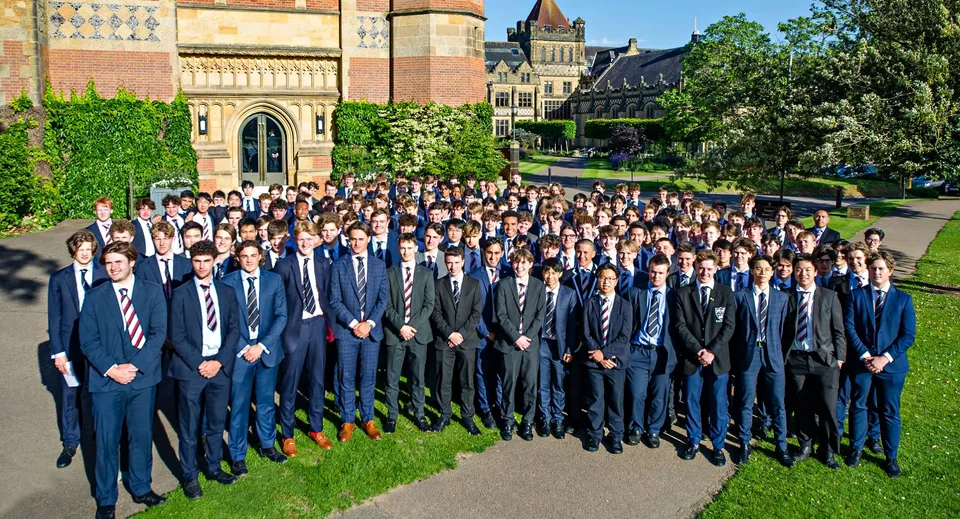 Un grande gruppo di studenti e studentesse della Tonbridge School si trova in abito formale davanti a un edificio storico dalla ricca architettura e circondato da una verde vegetazione.