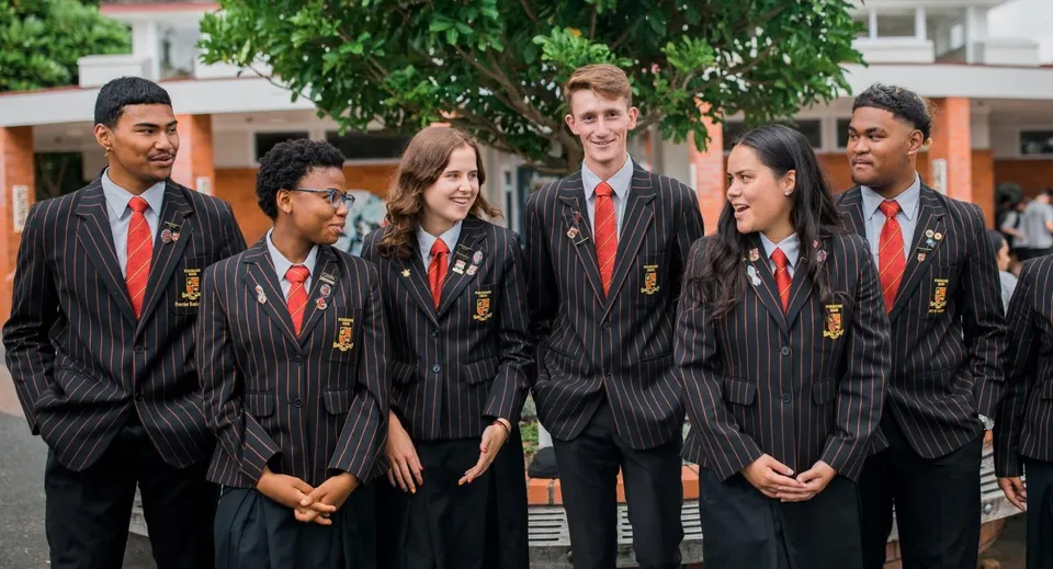 Un gruppo di studenti in toghe nere e cravatte rosse si trova davanti ad alberi verdi nel campus della Pukekohe High School.