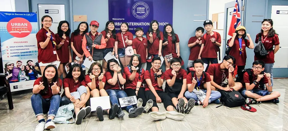 Un gruppo di studenti in uniforme rossa posa davanti a uno sfondo AIESEC International nel campus della Urban International High School.