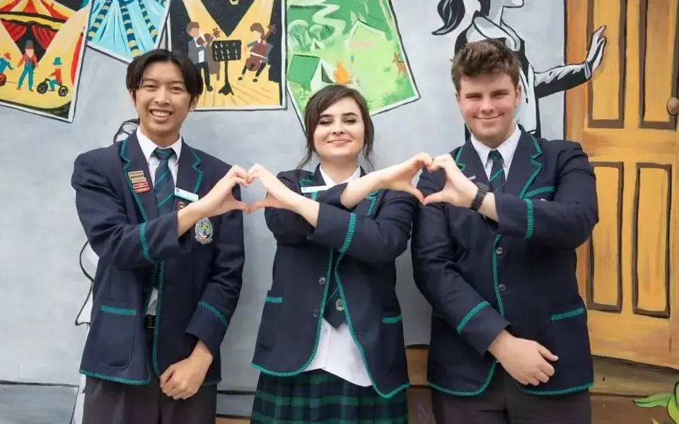 Tre studenti della Georgiana Molloy Anglican School in divisa scolastica formano dei cuori con le mani davanti a un murale.