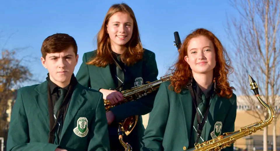 Tre studenti della Oak Bay Secondary High School in divisa verde sono in piedi con un sassofono e una tromba davanti a un cielo blu e alberi spogli.