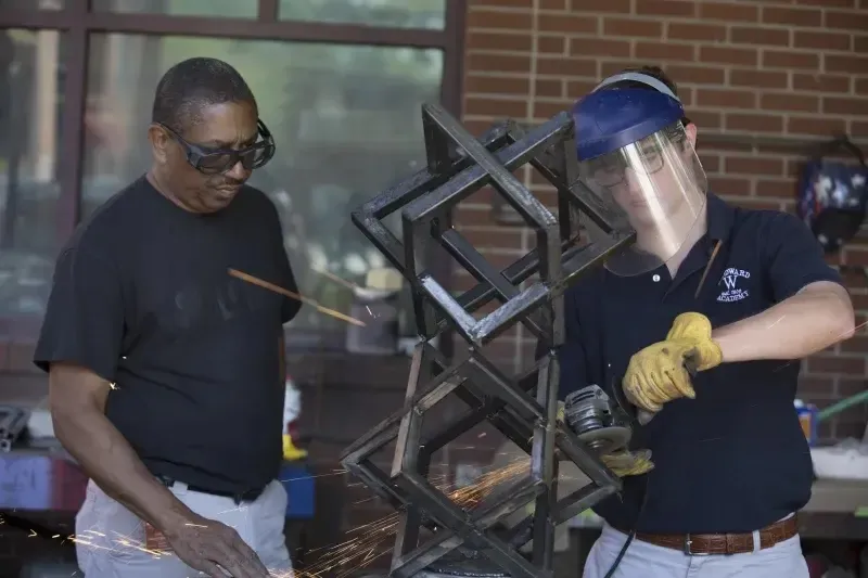 Dos personas trabajan en un marco de metal frente a una pared de ladrillo en los terrenos de la Woodward Academy.