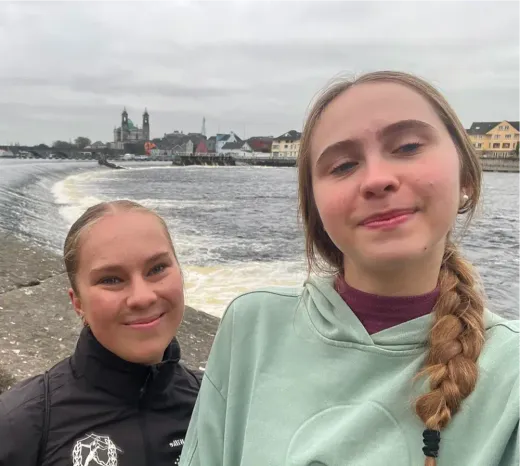 Lorena und eine Freundin machen ein Selfie am Fluss, im Hintergrund die Stadt in Irland
