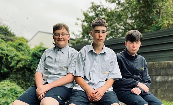Tres estudiantes con uniformes escolares están sentados juntos al aire libre en el recinto de la Rotorua Lakes High School ante un fondo verde.