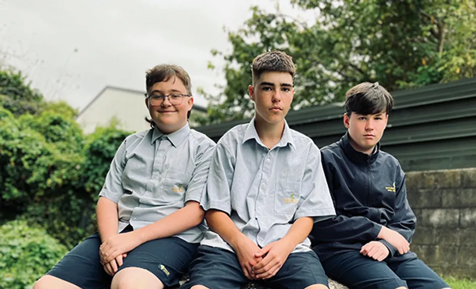 Tre studenti in uniforme scolastica seduti insieme all'aperto nel campus della Rotorua Lakes High School davanti a uno sfondo verde.