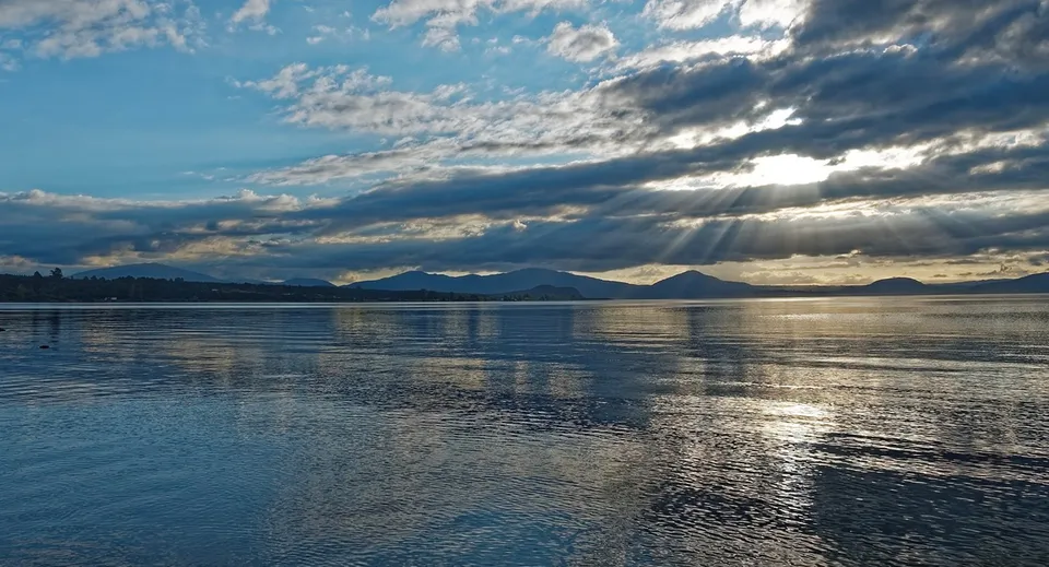 Un lago tranquillo con il cielo che si riflette e raggi di sole davanti alle montagne, vicino al Taupo-nui-a-Tia College.