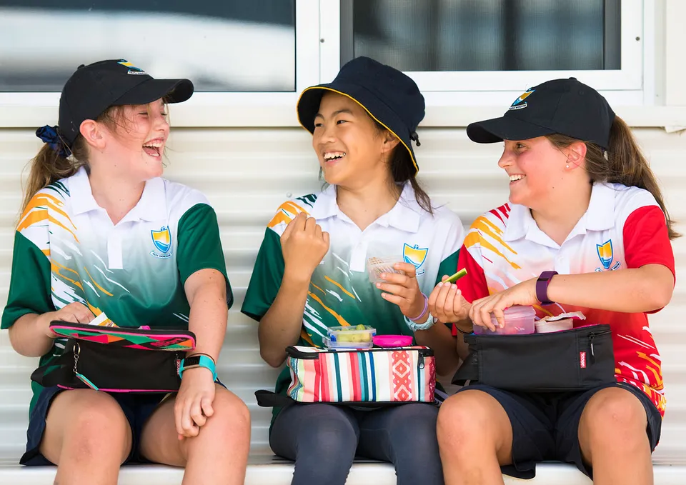 Tre studentesse della Esperance Anglican Community School siedono vicine, ridono e mangiano dai loro portapranzo.