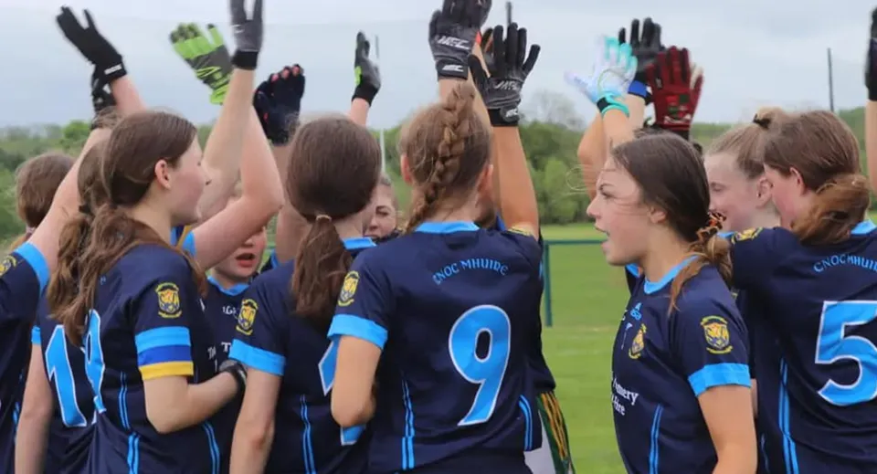 Un gruppo di giovani calciatrici in maglie blu e bianche festeggia insieme sul campo sportivo del Coláiste Mhuire (Ballygar).