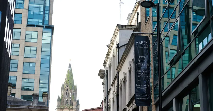 L'edificio moderno in vetro e acciaio dell'Abbey College a Manchester con l'alta guglia di un campanile sullo sfondo.