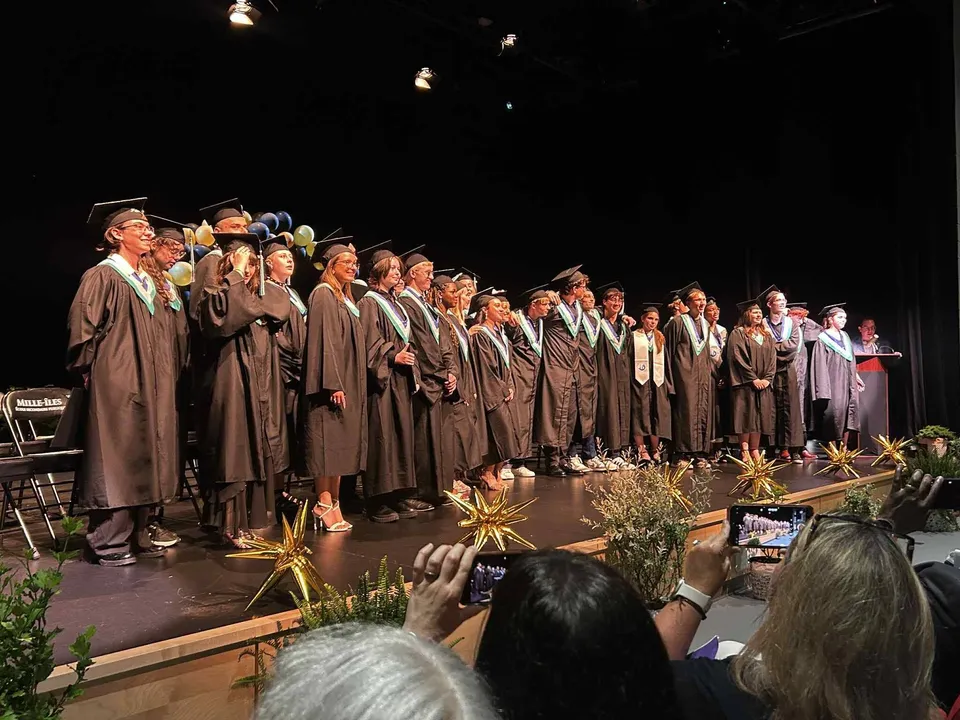 Un gruppo di diplomati della École secondaire publique Mille-îles si trova sul palco in abiti da cerimonia davanti a piante decorative.