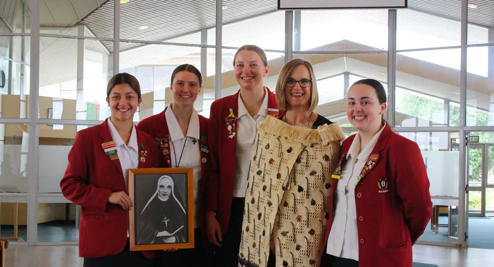 Cinque studentesse in toga rossa in una stanza luminosa con vetrate nel campus del Sacred Heart Girls' College a New Plymouth, una tiene in mano un ritratto incorniciato.