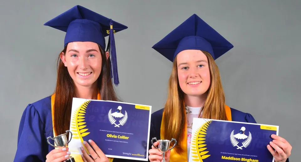 Due studentesse della Taumarunui High School indossano l'abito da diploma e tengono in mano i loro diplomi su uno sfondo grigio.