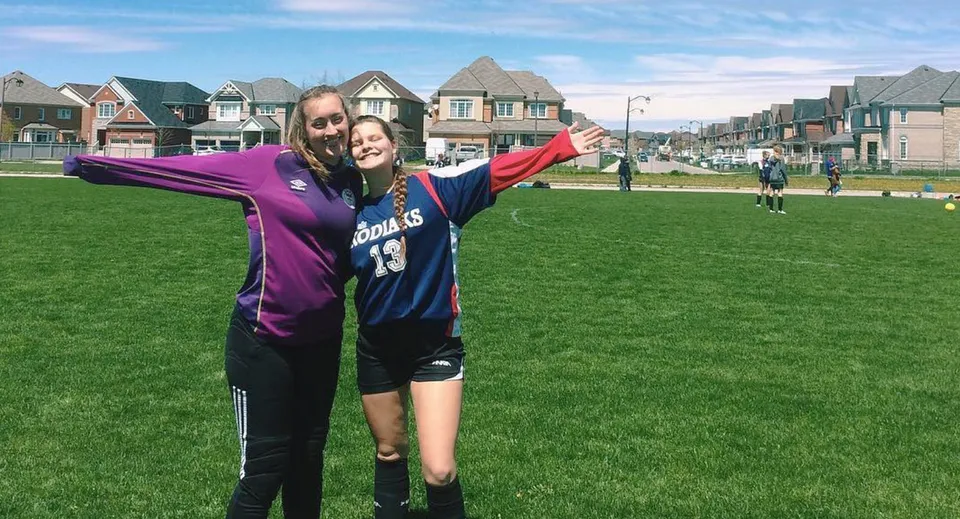 Due studentesse della Bradford District High School si abbracciano su un campo sportivo davanti a case colorate.
