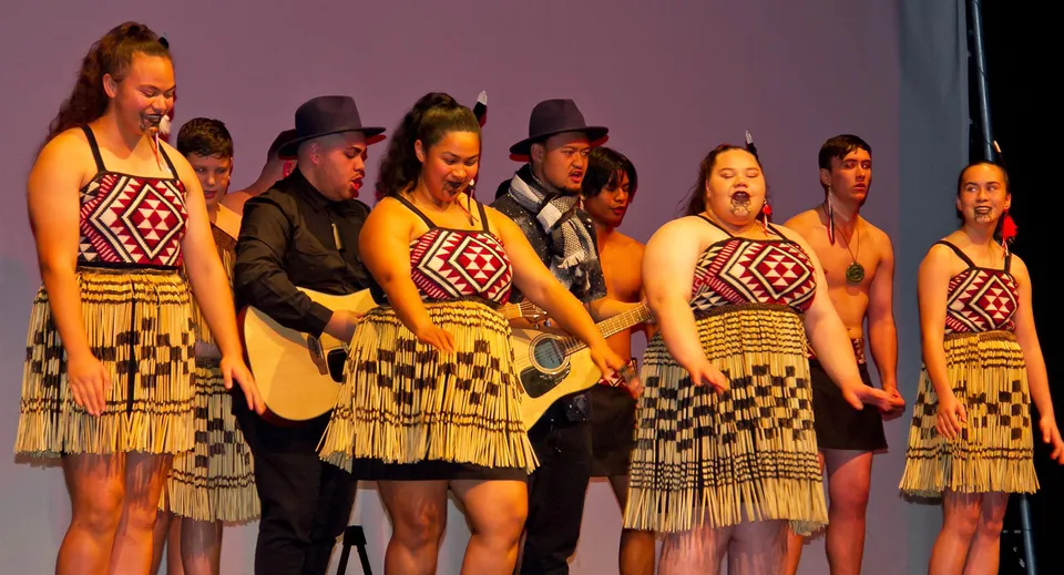 Studenti del Glenfield College eseguono danze indigene tradizionali in costumi tradizionali su un palco con uno sfondo viola.