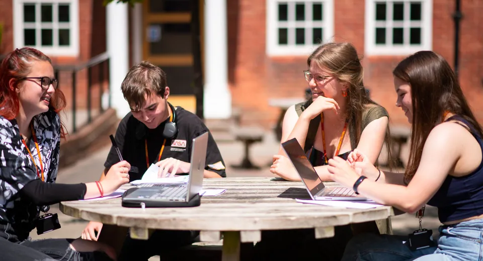 Studenti del College of Richard Collyer discutono all'aperto attorno a un tavolo di legno con i laptop.