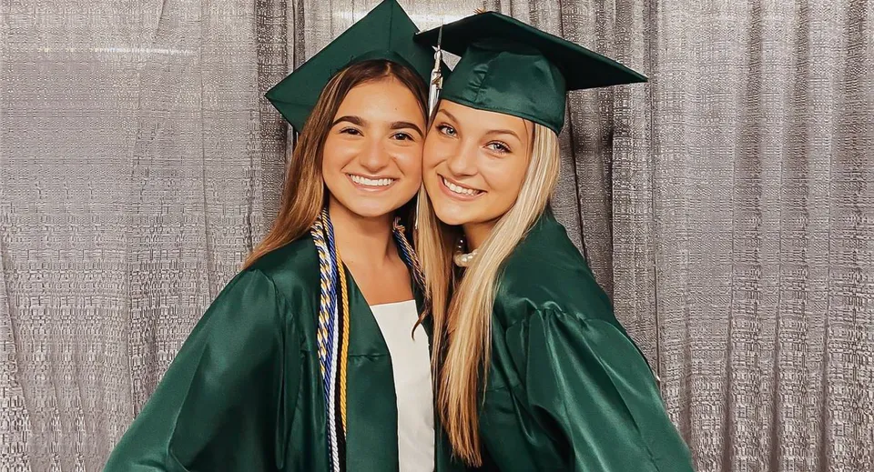 Due studentesse della Mercyhurst Prep School con la toga di diploma verde posano davanti a una tenda grigia.