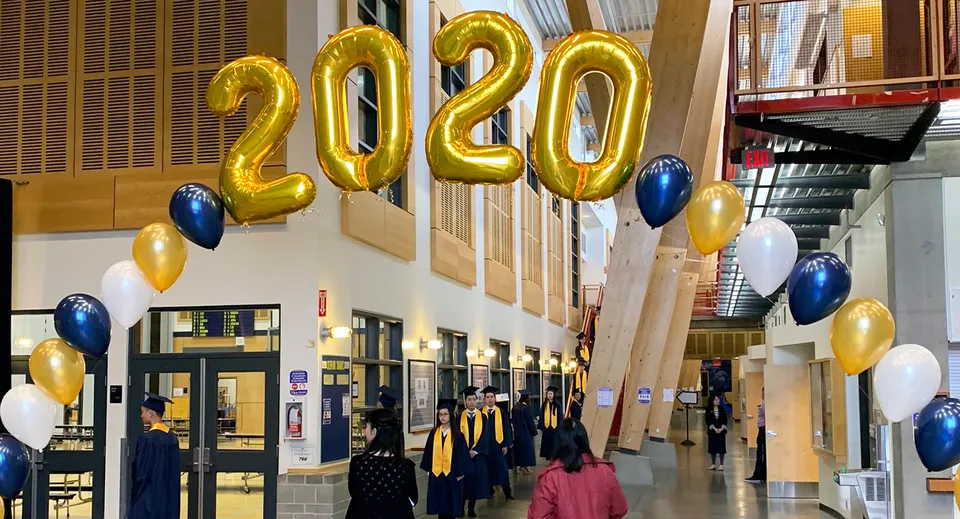 Studenti e studentesse della Steveston-London Secondary School in toga da diploma sotto grandi palloncini dorati del 2020 in un salone.