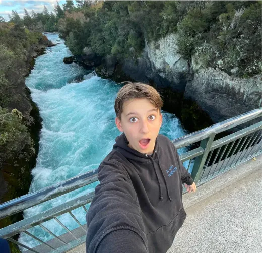 Junge macht ein Selfie auf einer Brücke über einen türkisen Fluss mit Hochwasser in Neuseeland