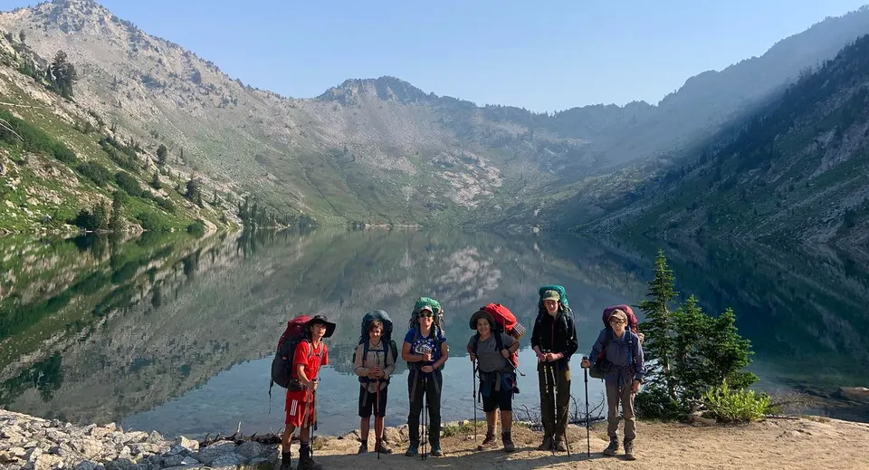 Un gruppo di studenti si trova sulla riva rocciosa di un lago davanti ad alte montagne sul terreno della Riverstone International School.