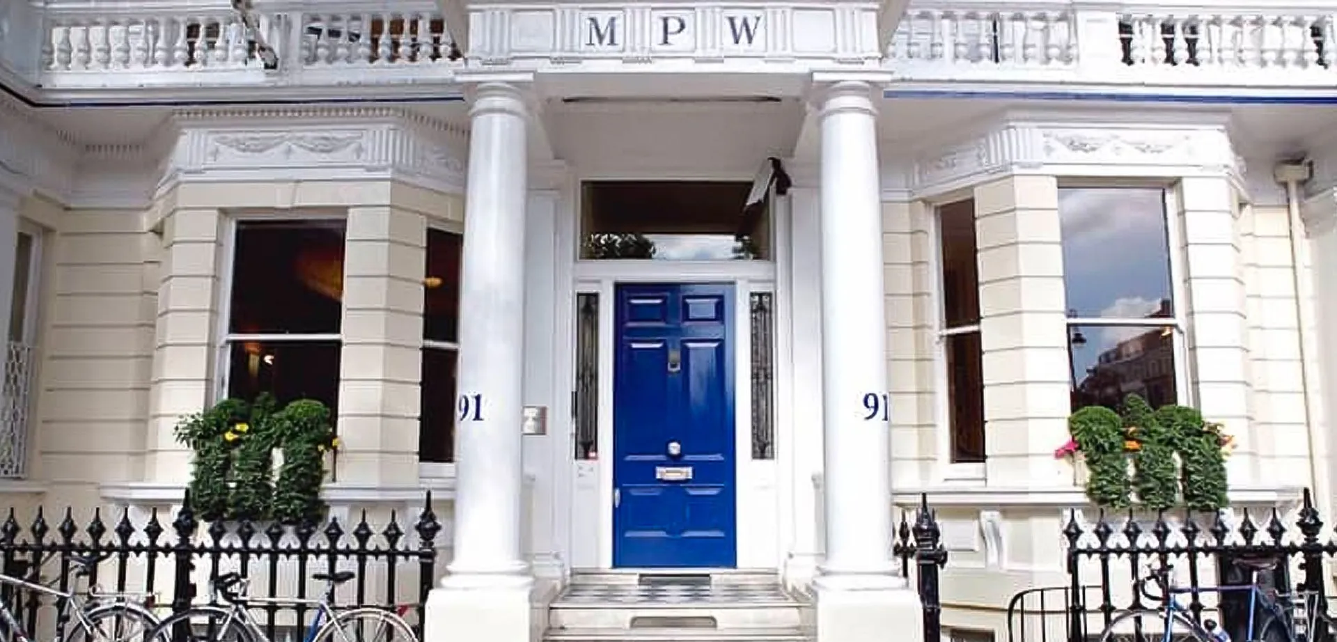 L'edificio bianco con colonne bianche e una porta d'ingresso blu nel campus della MPW London è circondato da piante verdi.