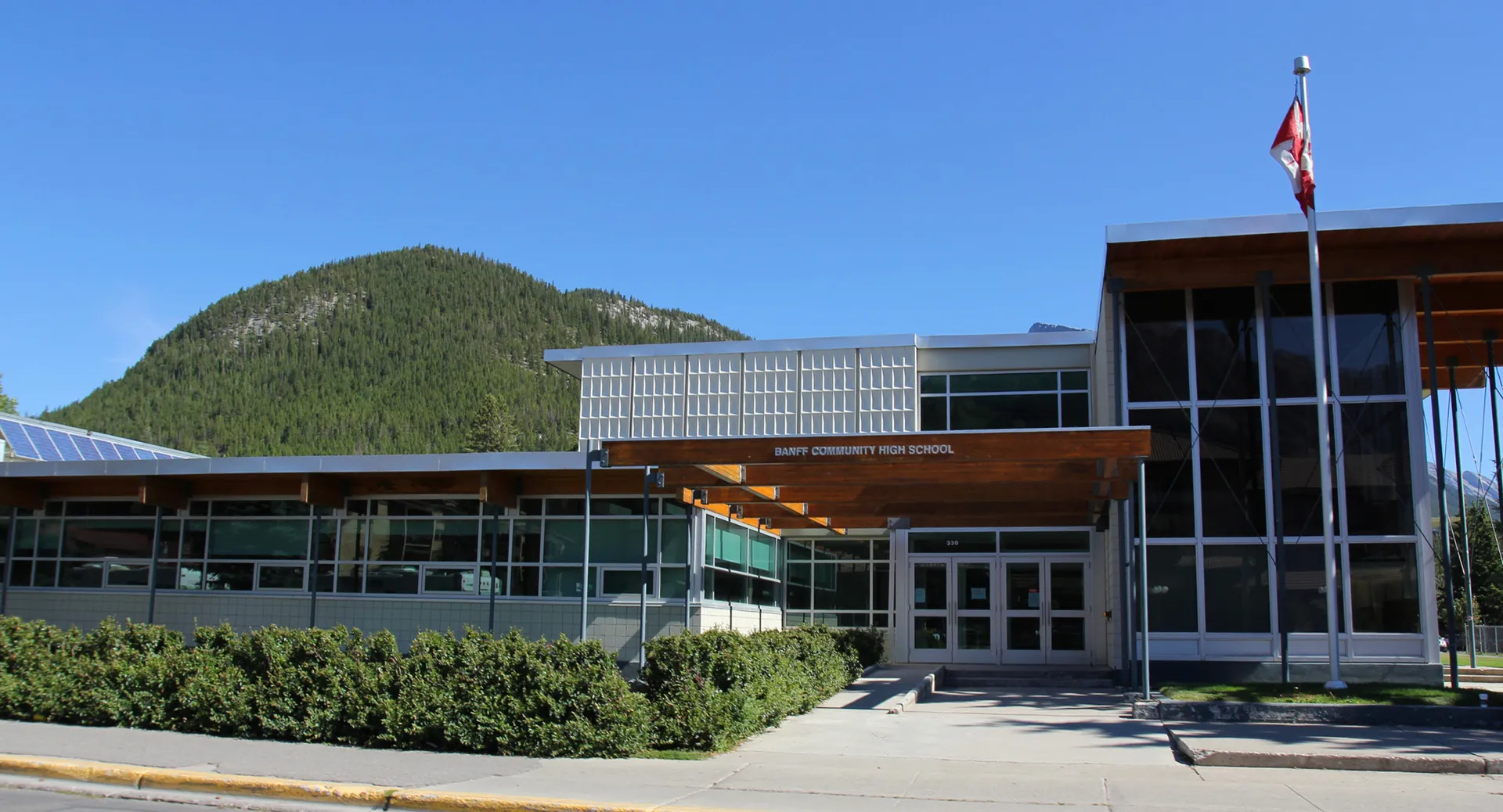 Edificio moderno della Banff Community High School con bandiera canadese davanti a una montagna boscosa.
