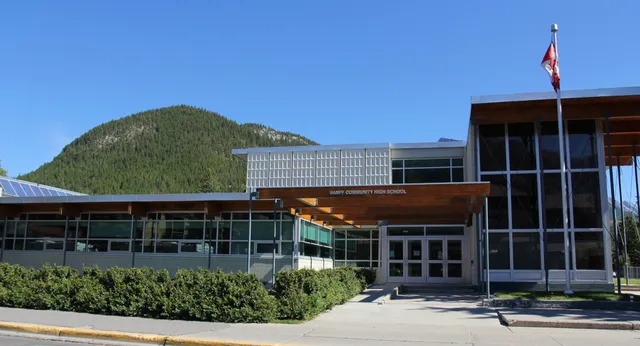 Edificio moderno de la Banff Community High School con la bandera canadiense frente a una montaña boscosa.
