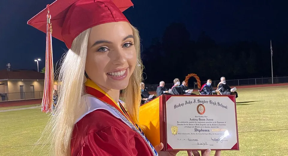 Una giovane donna con toga rossa e tocco tiene in mano il proprio diploma di notte sul campo da calcio della Bishop J Snyder High School.