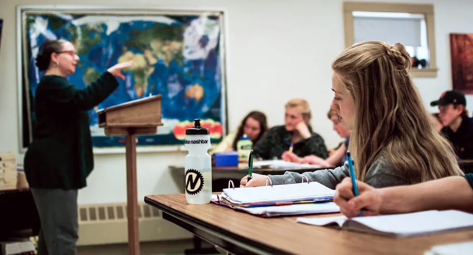 Gli studenti della George Stevens Academy prendono appunti in classe mentre una professoressa parla davanti a una carta geografica.