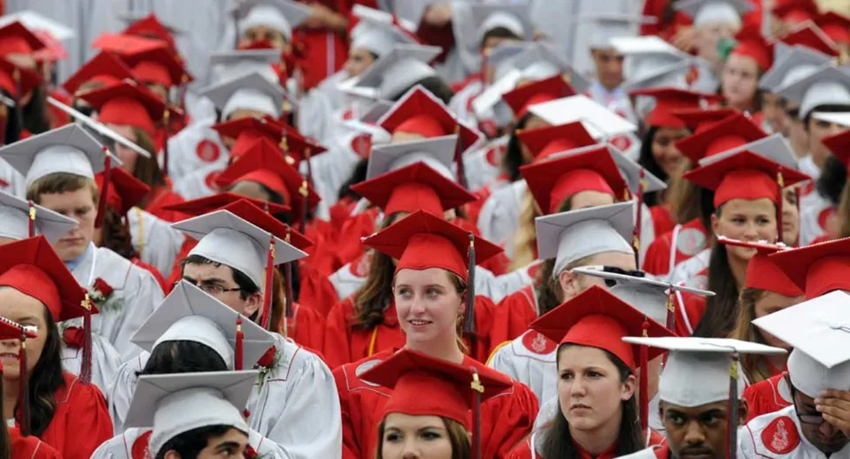 Un grande gruppo di studenti della Gloucester High School indossa toge e tocchi rossi per la cerimonia di diploma su uno sfondo neutro.