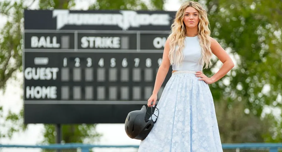 Una giovane donna sta in piedi davanti al tabellone segnapunti di una partita di baseball nel campus del Bonneville Joint School District e tiene in mano un cappello nero.