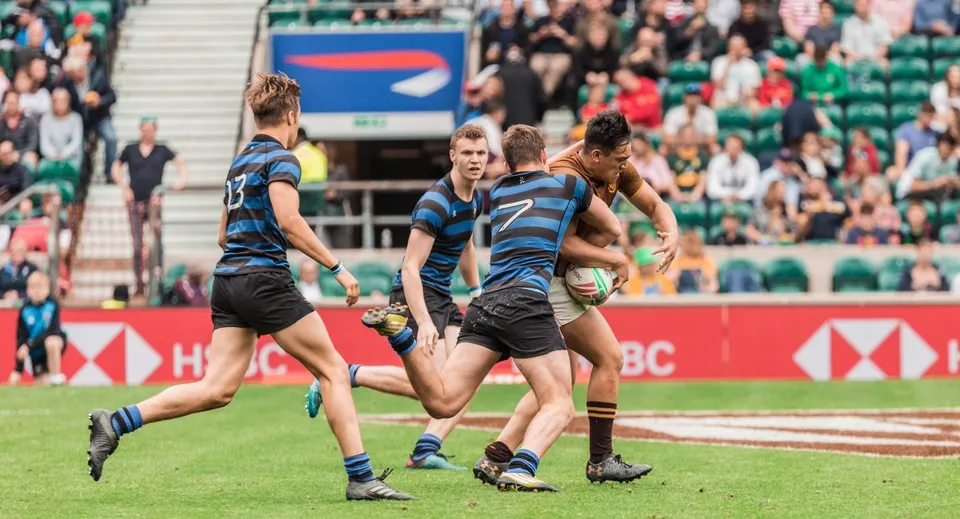 Studenti del Dulwich College giocano una partita di rugby su un campo d'erba con spettatori sullo sfondo.