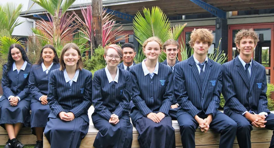 Un gruppo di studenti in divisa scolastica si trova davanti all'edificio del Birkenhead College con piante tropicali sullo sfondo.