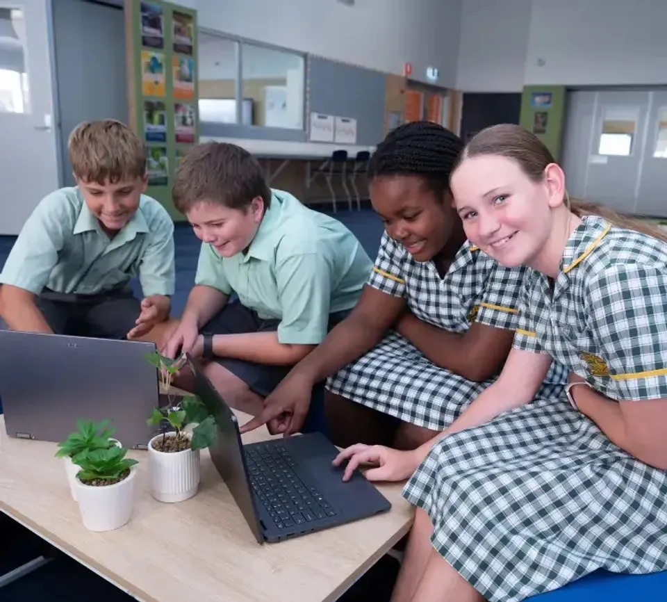 Quattro studenti della Frederick Irwin Anglican School in divisa scolastica siedono a un tavolo con laptop e piccole piante.