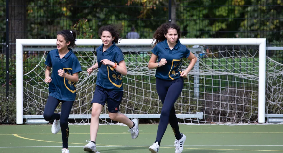 Tre studentesse della Kensington Park School corrono sul campo sportivo davanti a un palo della porta.