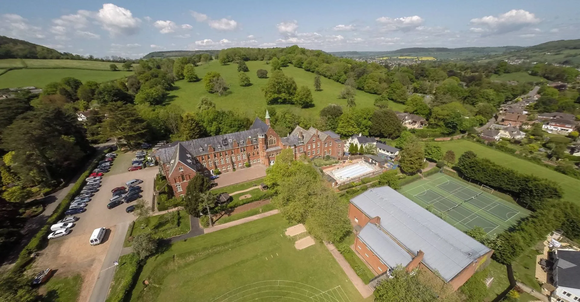 L'edificio storico della St. John’s School Sidmouth è immerso tra verdi colline e paesaggi boschivi, con un campo da tennis in primo piano.