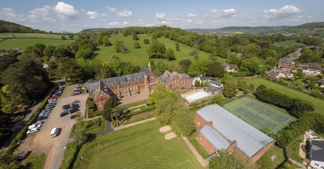 L'edificio storico della St. John’s School Sidmouth è immerso tra verdi colline e paesaggi boschivi, con un campo da tennis in primo piano.