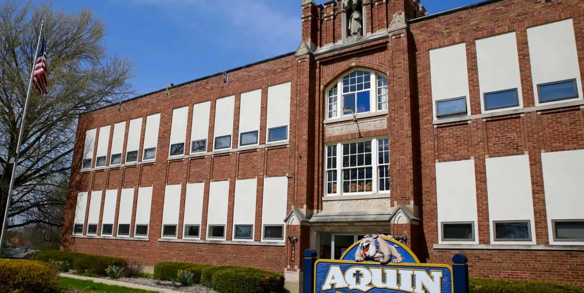 L'edificio in mattoni a più piani della Aquin High School è circondato da alberi e mostra una bandiera americana.
