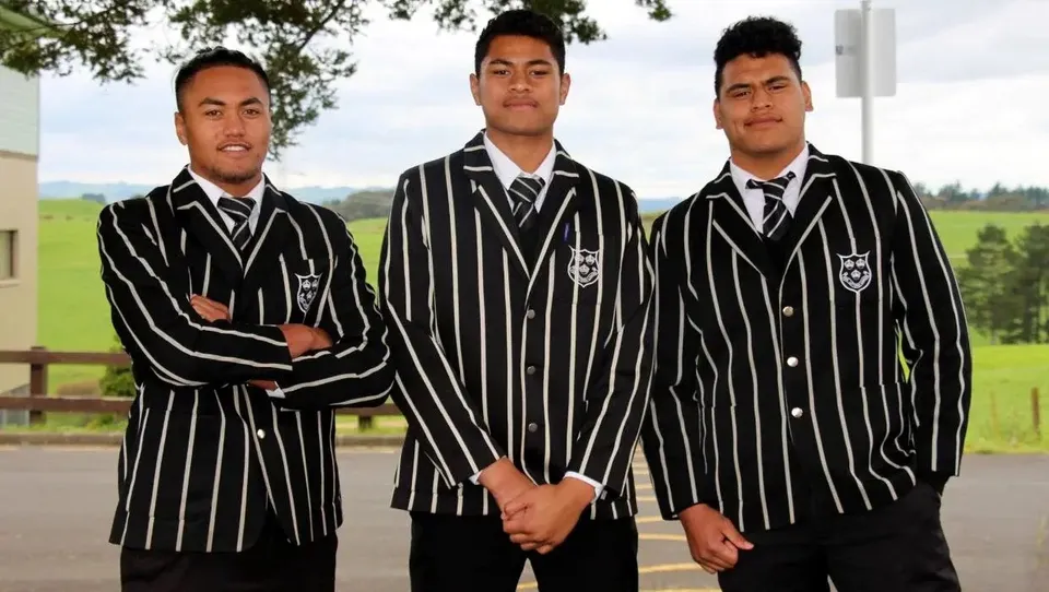 Tre studenti del Wesley College sono in piedi all'aperto su un prato davanti agli alberi, in eleganti abiti formali bianchi e neri.