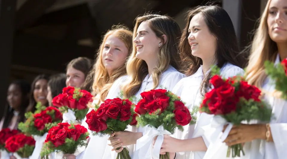 Un gruppo di giovani donne in abito bianco tiene delle rose rosse davanti a uno sfondo scuro nel campus della Santa Catalina School.