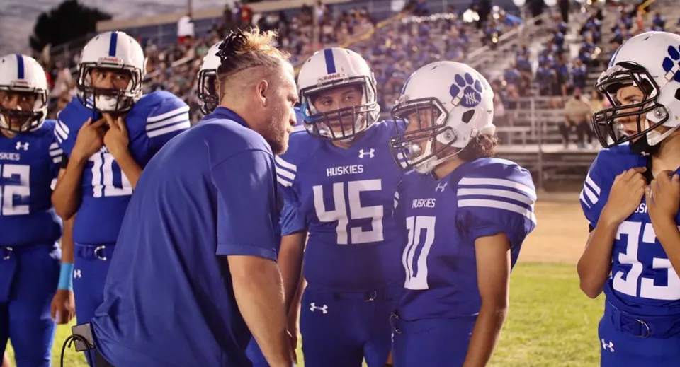 Un gruppo di giocatori di football in uniformi blu si trova sul campo da gioco del Los Angeles Unified School District con spettatori sullo sfondo.
