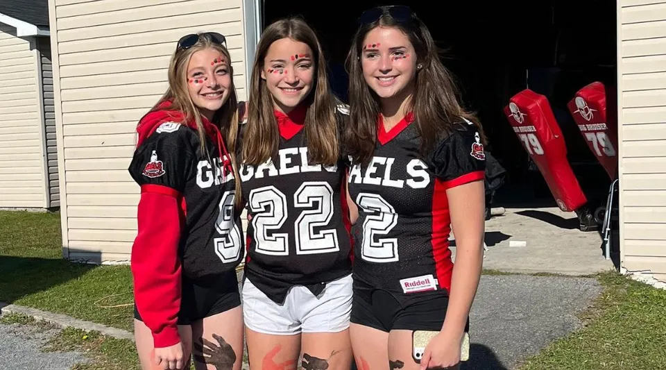Tre studentesse della Glengarry District High School posano con divise sportive uguali davanti a un edificio in legno.