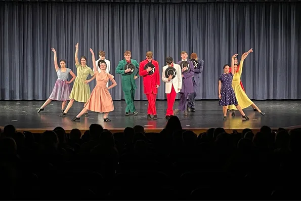 Grupo de estudiantes actuando en un escenario de teatro vistiendo trajes y vestidos de colores