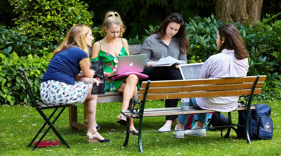 Studentesse dell'Exeter College sono sedute su una panchina nell'area verde all'aperto e studiano insieme con laptop e libri.