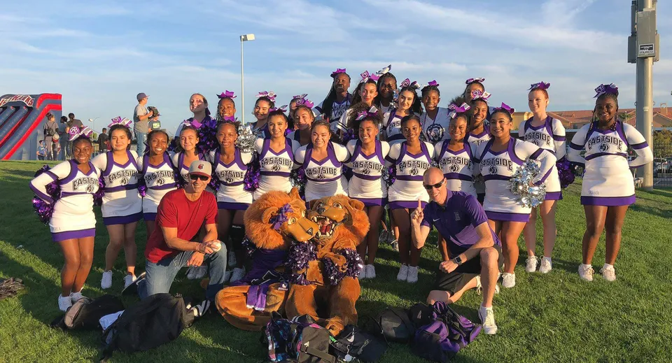 Un gruppo di cheerleader in uniforme viola e bianca sta sul campo sportivo dell'Antelope Valley Union High School District davanti a un cielo azzurro.