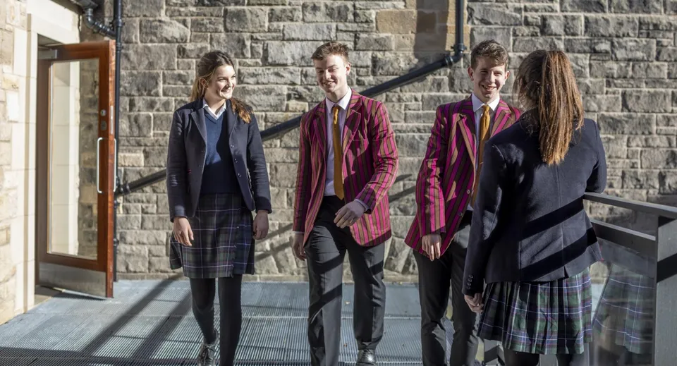 Quattro persone indossano un abbigliamento formale e sono in piedi davanti a un edificio in pietra nel campus del Fettes College.