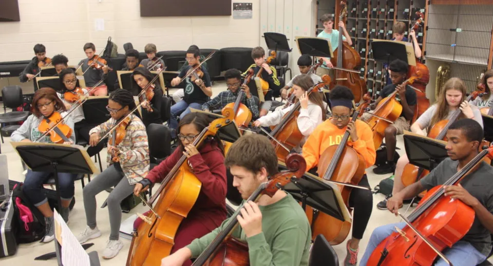 Un grande gruppo di studenti del Columba College suona strumenti a arco insieme in una sala da concerto.
