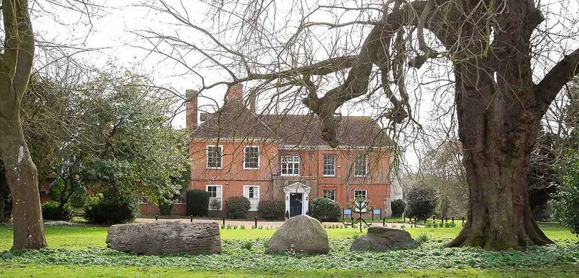 Un edificio storico in mattoni con ingresso a colonnato nel campus del Stoke College circondato da alberi spogli e grandi pietre.